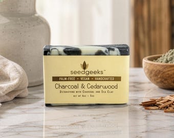 May include: A rectangular bar of soap with a black and white marbled pattern, wrapped in a beige paper label. The label reads seedgeeks, palm-free, vegan, handcrafted, Charcoal & Cedarwood, detoxifying with charcoal and sea clay, net wt 4oz.