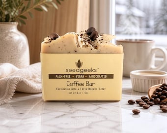 May include: A rectangular bar of soap topped with whole coffee beans and grounds. The beige paper label reads seedgeeks, Palm-Free, Vegan, Handcrafted, Coffee Bar, Exfoliating with a Fresh Brewed Scent, and Net Wt 4oz - 5oz in black text.