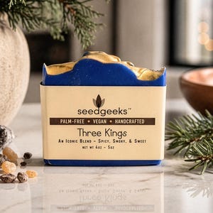 May include: A rectangular bar of blue soap with a gold-colored top, wrapped in a beige paper label. The label reads seedgeeks, Palm-Free, Vegan, Handcrafted, Three Kings, An Iconic Blend - Spicy, Smoky, & Sweet, and Net Wt 4oz - 5oz.