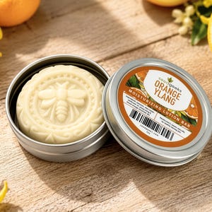 K&ouml;nnte beinhalten: Ein runder, cremefarbener Feuchtigkeits-Lotion-Riegel mit eingepr&auml;gtem Bienenmotiv in einer runden silbernen Metalldose. Der Deckel tr&auml;gt ein Etikett mit dem Text "seedgeeks ORANGE YLANG MOISTURIZING LOTION BAR" sowie einen Strichcode.