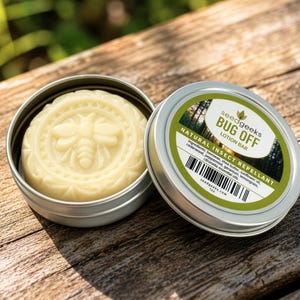 May include: A round, off-white lotion bar embossed with a bee design, housed in a silver metal tin. The lid features a green and white label with the text "seedgeeks BUG OFF LOTION BAR NATURAL INSECT REPELLANT" and a list of ingredients.