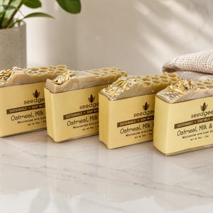 May include: Four rectangular bars of soap with a honeycomb-textured top, topped with oat flakes. Each bar is wrapped in a yellow paper label printed with the text 'seedgeeks', 'Sustainable Goat Milk', and 'Oatmeal, Milk & Honey', net wt 4oz - 5oz.