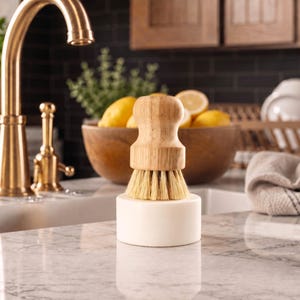 Puede incluir: Un cepillo de cocina con mango de madera y cerdas naturales, con una base blanca, sobre una encimera de m&aacute;rmol. Un grifo dorado y un cuenco con limones est&aacute;n en el fondo. El cepillo es para la cocina.