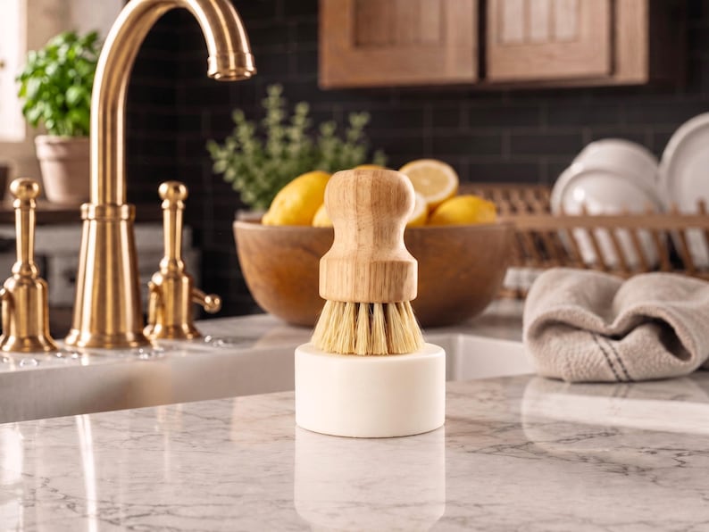 Puede incluir: Un cepillo de cocina con mango de madera y cerdas naturales, con una base blanca, sobre una encimera de m&aacute;rmol. Un grifo dorado y un cuenco con limones est&aacute;n en el fondo. El cepillo es para la cocina.