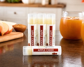 May include: Four cylindrical lip balm tubes with clear caps and white labels. Three tubes stand vertically, and one lies horizontally in front. Each label features the text seedgeeks APPLE CIDER Lip Balm and a small apple graphic.