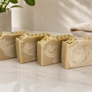 May include: Four rectangular bars of soap arranged in a row. Each bar features a light tan and cream marbled swirl pattern, a textured honeycomb design on the top edge, and a scattering of light brown oat flakes embedded into the top surface.