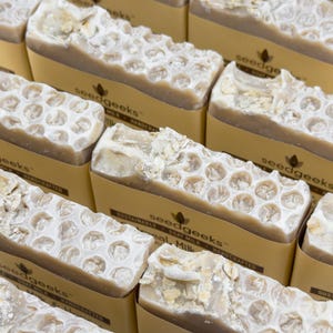 May include: Multiple rectangular bars of tan soap featuring a textured honeycomb pattern on the top surface and embedded oat flakes. Each bar is wrapped in a tan paper band printed with the brand name seedgeeks and the text sustainable goat milk.