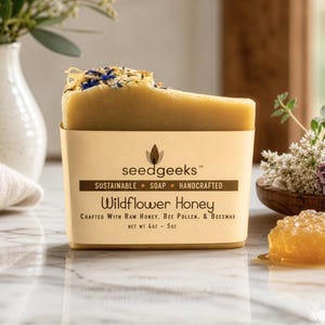May include: A bar of Wildflower Honey soap by seedgeeks, displayed in a beige paper sleeve. The soap is light brown with dried flower petals on top. The packaging includes the text "Wildflower Honey" and "Crafted With Raw Honey, Bee Pollen, & Beeswax".