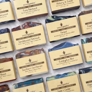 May include: An overhead shot of several bars of soap, each in a rectangular shape and wrapped in a tan paper label. The soaps have various colors and patterns, with labels indicating different scents like Charcoal & Cedarwood and Eucalyptus Mint. Each bar is approximately 4 oz.