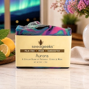 May include: A rectangular bar of soap with a marbled pattern of purple, pink, and teal colors. The soap is wrapped in a beige paper label that reads seedgeeks, palm-free, vegan, handcrafted, Aurora, a stellar blend of patchouli, citrus & musk, net wt 4oz - 5oz.