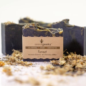 May include: Three dark gray soap bars with a brown label that reads "Forest" and "Activated Charcoal with Patchouli & Mint." The soap is surrounded by dried herbs. The label also says "All Natural, Vegan, Handcrafted." Each bar weighs 4.5 oz.