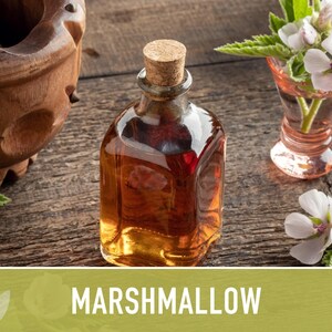 A bottle of amber-colored liquid, possibly a syrup or essential oil, sits on a wooden surface alongside a glass vase filled with white flowers and a wooden mortar and pestle.