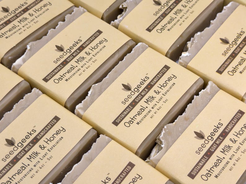 May include: Multiple rectangular bars of soap wrapped in tan paper labels. The labels feature the text seedgeeks, Sustainable, Goat Milk, Handcrafted, Oatmeal, Milk & Honey, Moisturizing with Light Exfoliation, and Net Wt 4oz - 5oz.
