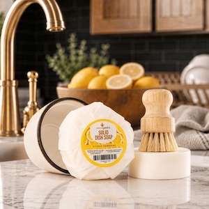 Puede incluir: Una pastilla de jab&oacute;n s&oacute;lido para lavar platos con aroma c&iacute;trico, un plato de cer&aacute;mica blanco y un cepillo para platos de madera se exhiben sobre una encimera de m&aacute;rmol. El jab&oacute;n est&aacute; etiquetado como "Citrus Sunshine". Un grifo dorado est&aacute; en el fondo.