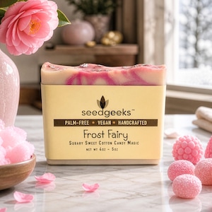 May include: A bar of soap with a light yellow label featuring the text "seedgeeks" and "Frost Fairy". The soap has a pink and white swirl design. Decorative pink candies and flowers are arranged around the soap.