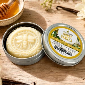 May include: A round, off-white moisturizing lotion bar embossed with a bee design, housed in a silver metal tin. The lid features a yellow and white label with the text "seedgeeks HONEY VANILLA MOISTURIZING LOTION BAR" and a barcode.