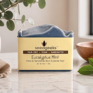 May include: A rectangular bar of dark blue soap wrapped in a beige paper label. The label reads seedgeeks, Palm-Free, Vegan, Handcrafted, Eucalyptus Mint, Fresh & Invigorating Daily Cleansing Soap, and Net Wt 4oz - 5oz in black text.