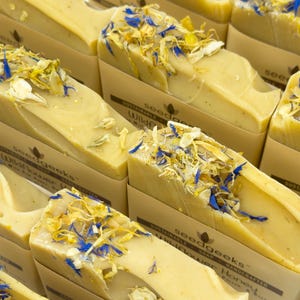 May include: Multiple rectangular bars of light yellow soap topped with dried blue and yellow flower petals. Each bar is wrapped in a beige paper band featuring the text seedgeeks, sustainable soap, and wildflower honey in dark brown ink.