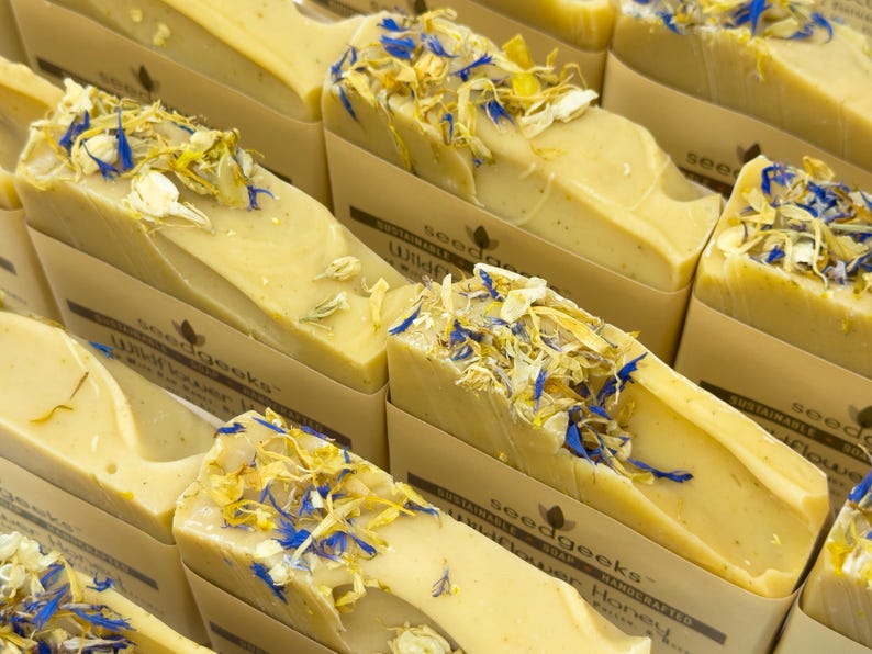 May include: Multiple rectangular bars of light yellow soap topped with dried blue and yellow flower petals. Each bar is wrapped in a beige paper band featuring the text seedgeeks, sustainable soap, and wildflower honey in dark brown ink.