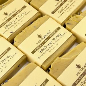 May include: Multiple rectangular bars of Wildflower Honey soap by seedgeeks. Each bar is wrapped in a beige paper band featuring the brand name, the text Wildflower Honey, and the ingredients raw honey, bee pollen, and beeswax. Net wt 4oz - 5oz.