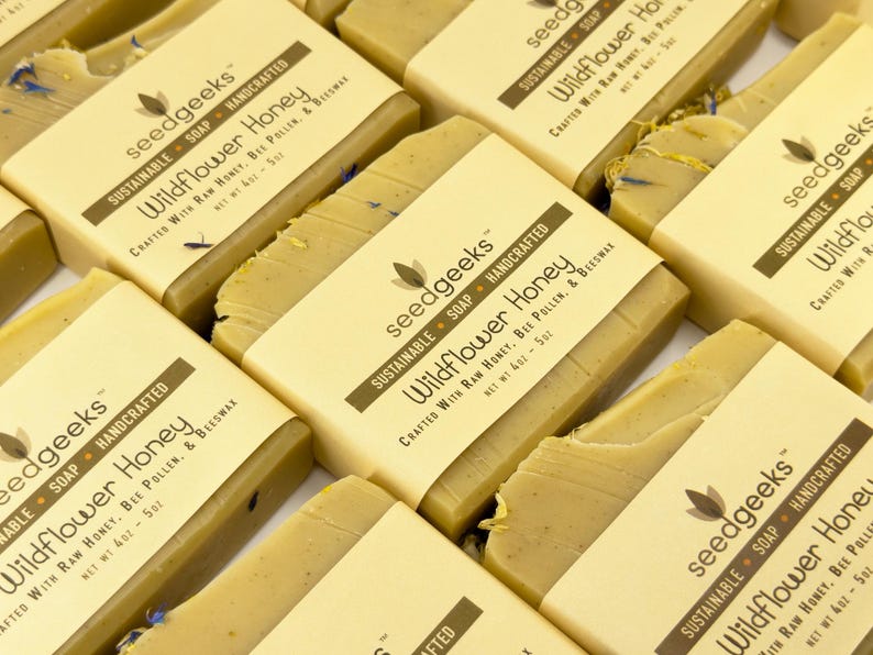 May include: Multiple rectangular bars of Wildflower Honey soap by seedgeeks. Each bar is wrapped in a beige paper band featuring the brand name, the text Wildflower Honey, and the ingredients raw honey, bee pollen, and beeswax. Net wt 4oz - 5oz.