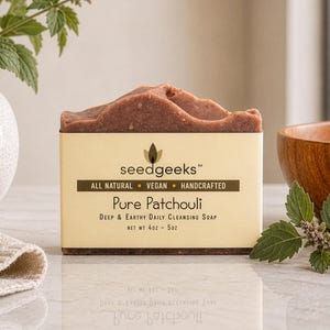 May include: A bar of Pure Patchouli soap with a textured, brown surface, displayed on a light-colored surface. The soap is labeled with the text "seedgeeks" and "Pure Patchouli". The image includes a wooden bowl, a vase with greenery, and a sprig of leaves.
