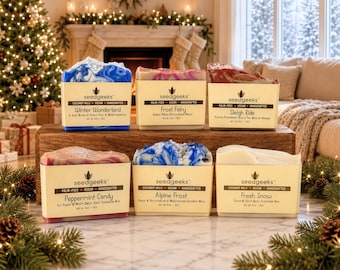 May include: Six rectangular bars of handcrafted soap arranged on a wooden block. Each bar features a cream-colored paper label with the brand name seedgeeks and specific scent names: Winter Wonderland, Frost Fairy, Sleigh Ride, Peppermint Candy, Alpine Frost, and Fresh Snow. The soaps display varied colors including blue, pink, red, and white swirls.