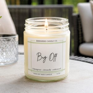 May include: A clear glass jar candle with a lit wick and cream-colored wax. The white label features the text Seedgeeks Candle Co., Bug Off, lemongrass citronella cedarwood, 100% soy wax candle, and 8 oz in black and green ink.