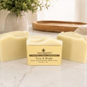 May include: Three rectangular bars of off-white soap. The center bar features a tan paper label with the text: seedgeeks, PALM-FREE, VEGAN, COCONUT MILK, Pure & Simple, NET WT 4oz - 5oz. The soap has a smooth, solid texture and a matte finish.