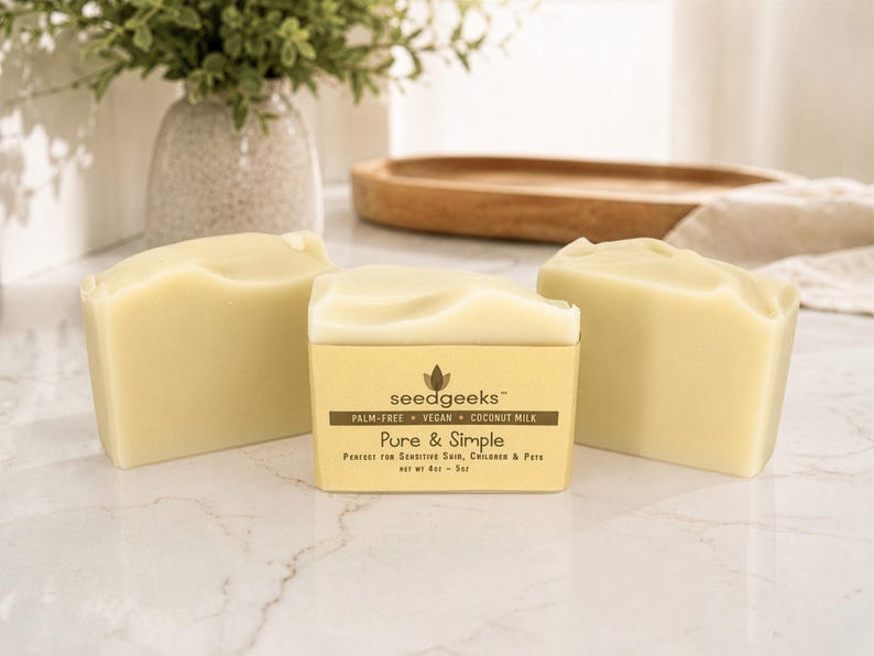 May include: Three rectangular bars of off-white soap. The center bar features a tan paper label with the text: seedgeeks, PALM-FREE, VEGAN, COCONUT MILK, Pure & Simple, NET WT 4oz - 5oz. The soap has a smooth, solid texture and a matte finish.