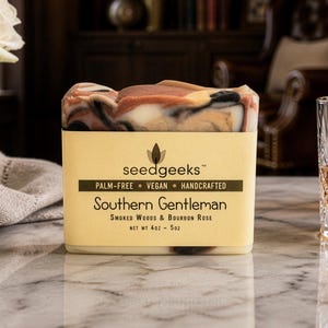 May include: A rectangular bar of soap with a marbled pattern of brown, black, and cream colours. The soap is wrapped in a beige paper label with the text seedgeeks, Palm-Free, Vegan, Handcrafted, Southern Gentleman, Smoked Woods & Bourbon Rose, Net Wt 4oz - 5oz.
