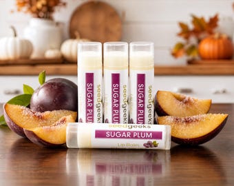 May include: Four cylindrical lip balm tubes labeled seedgeeks SUGAR PLUM. Three tubes stand upright, and one lies horizontally in front. The labels feature purple accents and a small plum graphic. Fresh whole and sliced plums are arranged nearby.