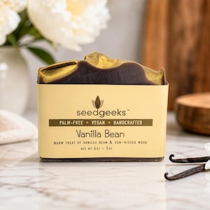 May include: A Vanilla Bean soap bar with a dark brown top and gold detailing, in a light yellow box. The box reads "seedgeeks" and "Palm-Free Vegan Handcrafted." The soap is displayed with vanilla pods and a white flower. Net weight is 113 grams.