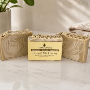 May include: Three rectangular bars of oatmeal, milk, and honey soap with a marbled beige and white pattern. The center bar features a gold label with the text seedgeeks, sustainable, goat milk, handcrafted, oatmeal, milk & honey, net wt 4oz - 5oz.