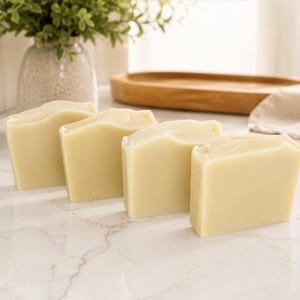 May include: Four rectangular bars of solid, off-white soap arranged in a staggered row. Each bar features a smooth, matte finish with slightly rounded top edges and a uniform, pale cream color throughout the entire surface of the soap blocks.