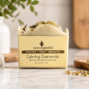 May include: A rectangular bar of off-white soap topped with dried chamomile flowers. The soap is wrapped in a beige paper label that reads seedgeeks, palm-free, vegan, handcrafted, Calming Chamomile, soothing with gentle exfoliation, net wt 4oz - 5oz.