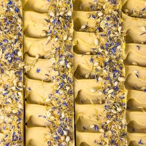 May include: Several rectangular bars of pale yellow soap arranged in a row. Each bar features a textured top surface decorated with a vertical strip of dried blue cornflower petals and small white flower buds embedded into the soap base.