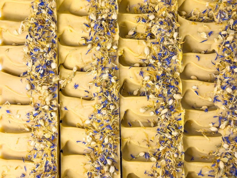 May include: Several rectangular bars of pale yellow soap arranged in a row. Each bar features a textured top surface decorated with a vertical strip of dried blue cornflower petals and small white flower buds embedded into the soap base.