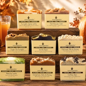 May include: Eight rectangular bars of handcrafted soap arranged on wooden blocks. Each bar features a beige paper label with the brand name seedgeeks and specific scent names including Pumpkin Spice Latte, Farmhouse Cider, Sugar Pie Pumpkin, Vanilla Bean, Oatmeal, Milk & Honey, Matcha Latte, Cinnamon, Oats & Honey, and Coffee Bar.