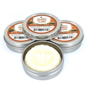 K&ouml;nnte beinhalten: Vier runde silberne Metalldosen mit feuchtigkeitsspendenden Lotionsriegeln. Drei Dosen sind geschlossen und mit Etiketten versehen, auf denen seedgeeks ORANGE YLANG MOISTURIZING LOTION BAR steht. Eine offene Dose zeigt einen festen wei&szlig;en Riegel.