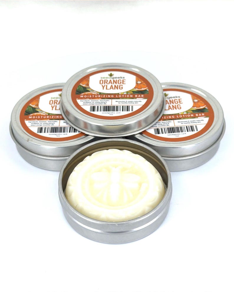K&ouml;nnte beinhalten: Vier runde silberne Metalldosen mit feuchtigkeitsspendenden Lotionsriegeln. Drei Dosen sind geschlossen und mit Etiketten versehen, auf denen seedgeeks ORANGE YLANG MOISTURIZING LOTION BAR steht. Eine offene Dose zeigt einen festen wei&szlig;en Riegel.