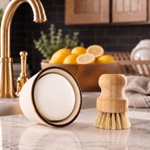 Puede incluir: Un jabonera crema con borde marr&oacute;n oscuro y un cepillo de cocina de madera con cerdas claras sobre una encimera de m&aacute;rmol. Un grifo dorado y limones en un cuenco de madera se ven al fondo.