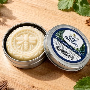 May include: A round, off-white moisturizing lotion bar embossed with a bee design, housed in a silver metal tin. The lid features a blue and white label with the text Cedar Patchouli, Moisturizing Lotion Bar, and a barcode.