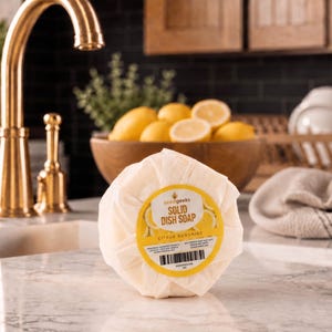 Puede incluir: Una pastilla de jab&oacute;n s&oacute;lido para lavar platos, redonda y envuelta, con una etiqueta amarilla y blanca. La etiqueta dice "Solid Dish Soap Citrus Sunshine" e incluye un c&oacute;digo de barras. El jab&oacute;n est&aacute; sobre una encimera de m&aacute;rmol cerca de un grifo dorado y un cuenco de madera con limones.