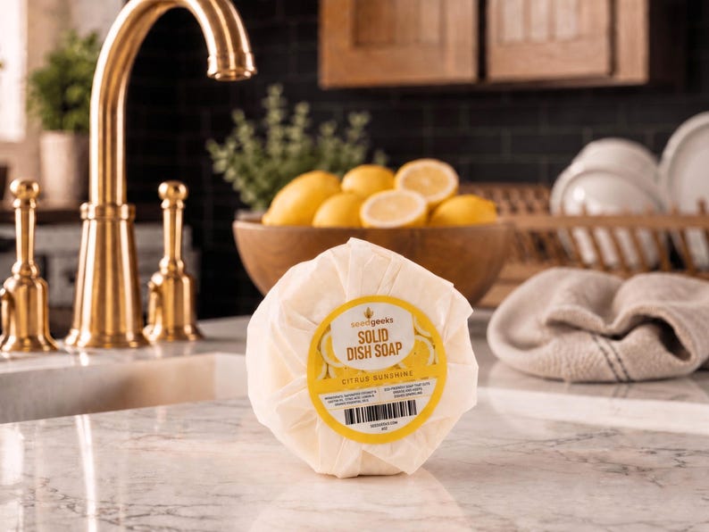 Puede incluir: Una pastilla de jab&oacute;n s&oacute;lido para lavar platos, redonda y envuelta, con una etiqueta amarilla y blanca. La etiqueta dice "Solid Dish Soap Citrus Sunshine" e incluye un c&oacute;digo de barras. El jab&oacute;n est&aacute; sobre una encimera de m&aacute;rmol cerca de un grifo dorado y un cuenco de madera con limones.