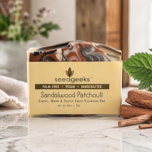 May include: A bar of handcrafted soap with a marbled design in shades of brown, white, and black, labeled "Sandalwood Patchouli." The soap is in a yellow box with the "seedgeeks" logo and text indicating it's palm-free, vegan, and a cleansing bar. The net weight is 4 oz - 5 oz.