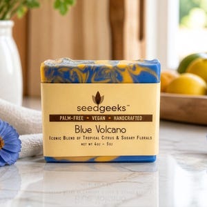 May include: A bar of "Blue Volcano" soap with blue and yellow marbling. The soap is wrapped in a light yellow label with the "seedgeeks" logo and text. The label states the soap is palm-free, vegan, and handcrafted, with a net weight of 4 oz - 5 oz.