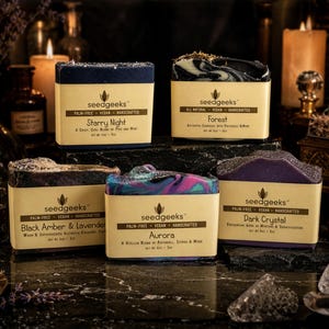 May include: Five rectangular bars of soap labeled seedgeeks, each with a tan paper band. The scents are Starry Night, Forest, Black Amber & Lavender, Aurora, and Dark Crystal. Each bar weighs 5 oz and features distinct colors and textures.
