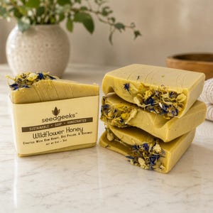 May include: A stack of three rectangular bars of pale yellow soap topped with dried blue and yellow wildflower petals. One bar is wrapped in a beige paper label reading seedgeeks Wildflower Honey Crafted With Raw Honey, Bee Pollen, & Beeswax Net Wt 4oz - 5oz.