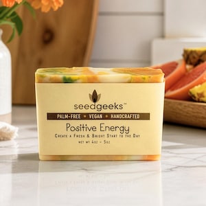 May include: A rectangular bar of soap with a marbled orange, yellow, and white pattern. The label reads seedgeeks, PALM-FREE, VEGAN, HANDCRAFTED, Positive Energy, CREATE A FRESH & BRIGHT START TO THE DAY, and NET WT 4OZ - 5OZ in dark brown text.